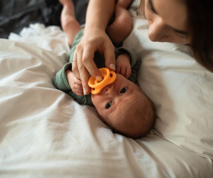 baby lying on a bed with his mom giving him an orange pacifier