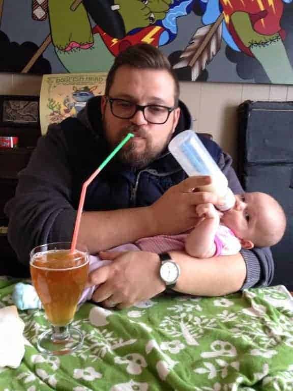 good father drinks beer through long straw while bottle feeding his baby